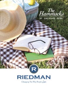 A picnic scene with a blanket, sunglasses on an open book, a straw hat, and a wicker basket on grass. Text reads: "The Hammocks at Orchard Park. Riedman. Changing The Way People Live.