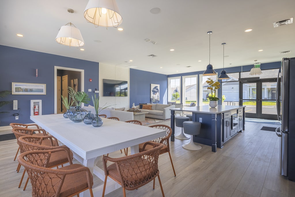 Open-concept living and dining area with wicker chairs, a long white dining table, modern kitchen, and large windows. Blue walls and hanging lights create a cozy atmosphere.