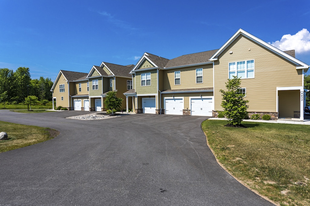 A row of modern, beige townhouses with attached garages, surrounded by a paved driveway and green lawns under a clear blue sky.