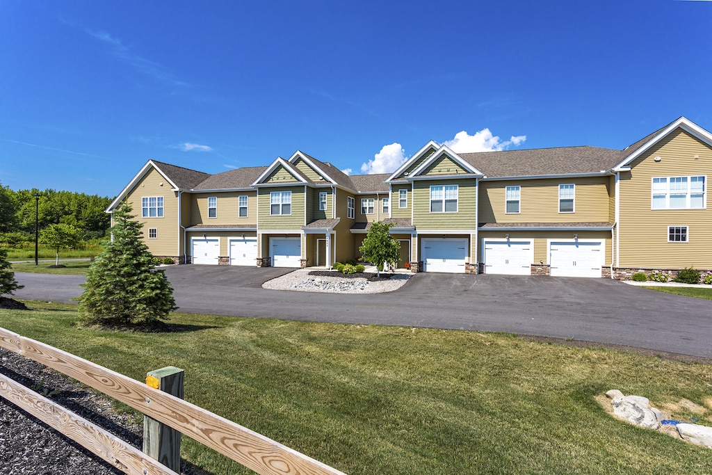 Two-story townhouse complex with beige siding and white garage doors, situated on a paved driveway, surrounded by grass, trees, and a wooden fence under a clear blue sky.