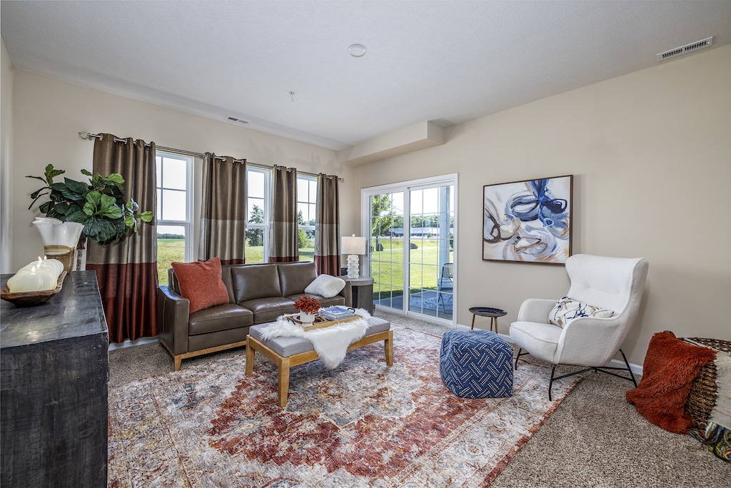 Living room with a sofa, armchair, and a coffee table on a patterned rug. Large windows and a sliding door overlook a grassy area. Wall art and decorative plants enhance the space.