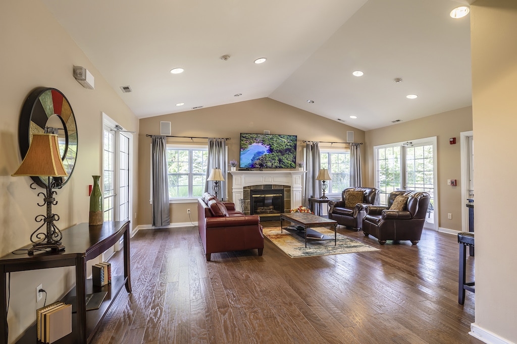 Spacious living room with hardwood floor, leather sofas, a coffee table, and a flat-screen TV above a fireplace. Large windows provide ample natural light.