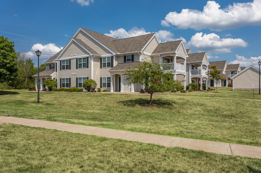 A two-story residential building with beige siding and a gabled roof is surrounded by a well-kept lawn and some trees, set against a partially cloudy blue sky.