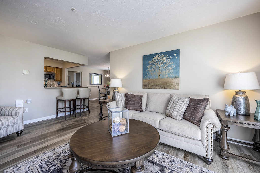 Living room with a beige sofa, wooden coffee table, two lamps, and a wall painting. Open view to a kitchen with bar stools and neutral-colored decor.