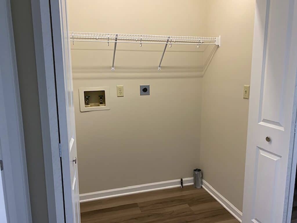 A small laundry room with light walls and wood flooring, featuring a white wire shelf and utility connections on the wall.