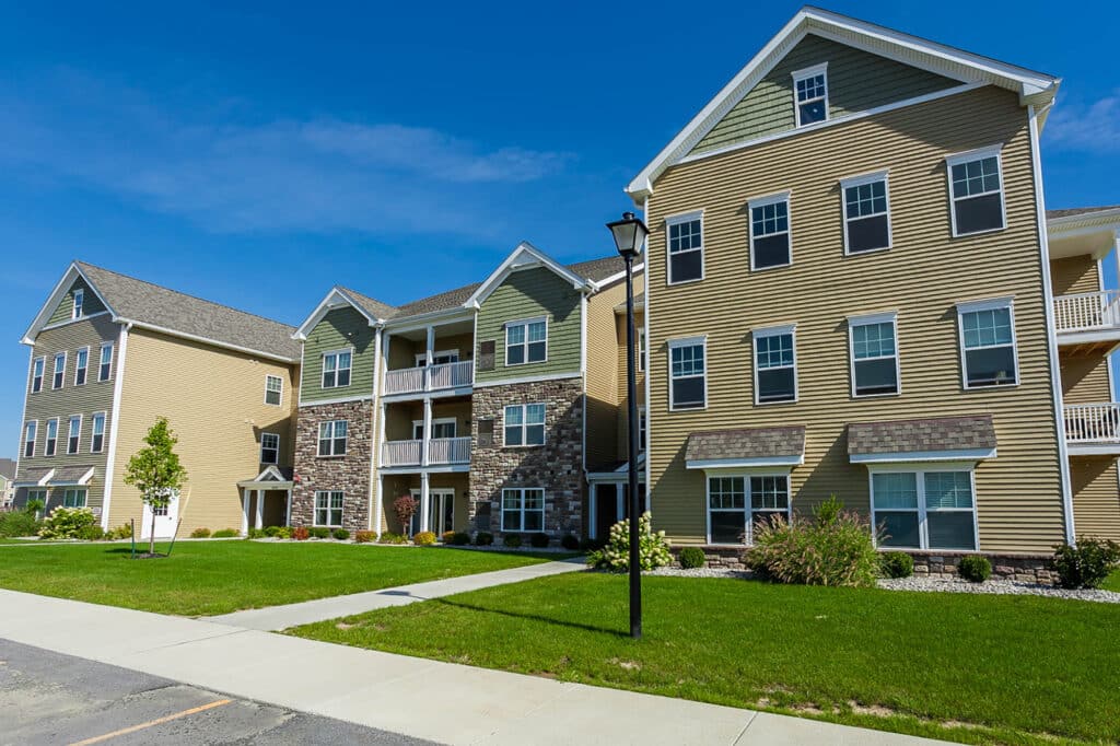 A modern apartment building with tan siding, large windows, and balconies, situated in a grassy area with a paved walkway.