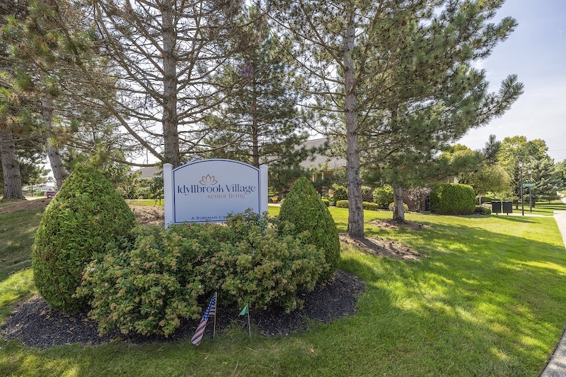 Sign for Idyllbrook Village Retirement Living surrounded by neatly trimmed bushes and tall trees in a sunny outdoor setting.