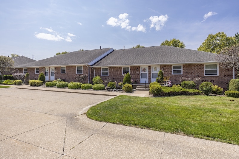 Row of brick single-story residential units with neatly trimmed bushes, trees, and a paved driveway under a clear blue sky.