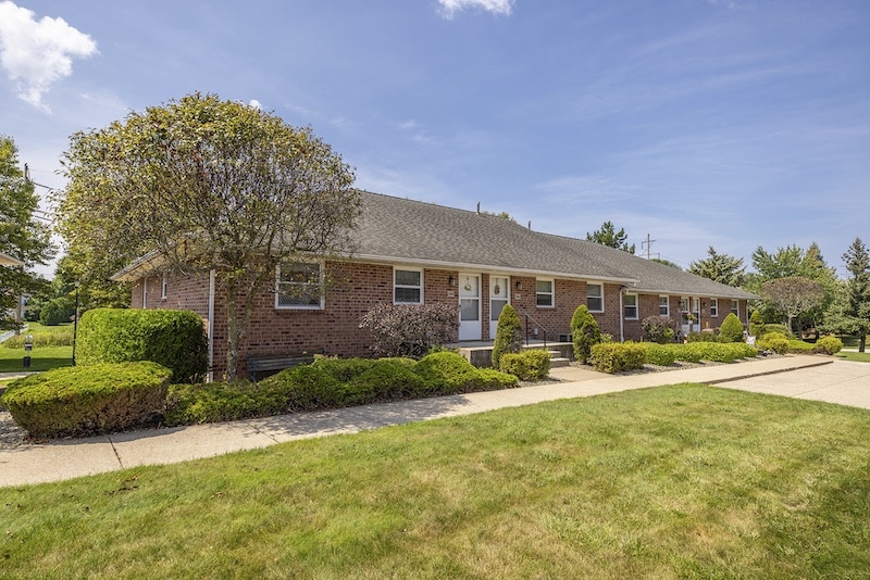 A single-story brick building with a gray roof is surrounded by neatly trimmed bushes and a grassy lawn under a clear blue sky.