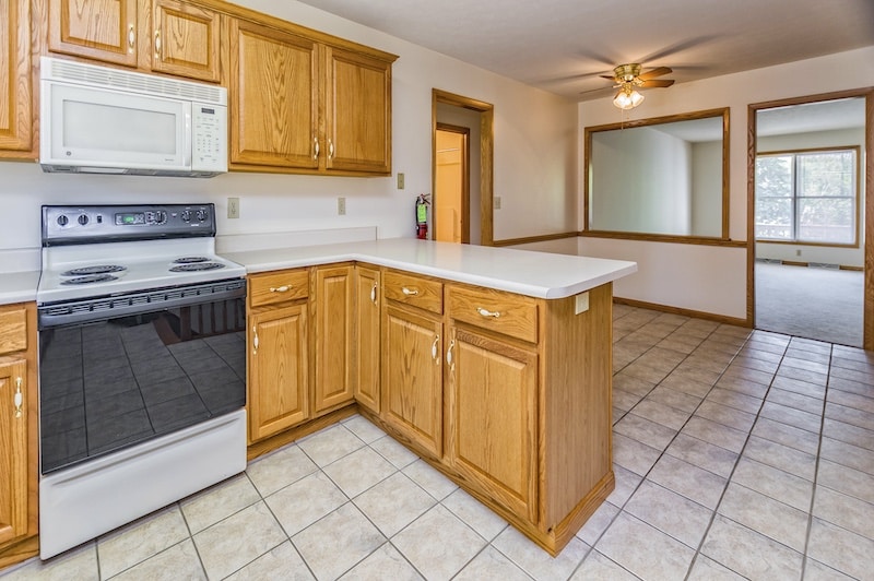 A kitchen with wood cabinets and white countertops features a white microwave, a black electric stove, and tiled flooring. A ceiling fan is in the adjacent open area.