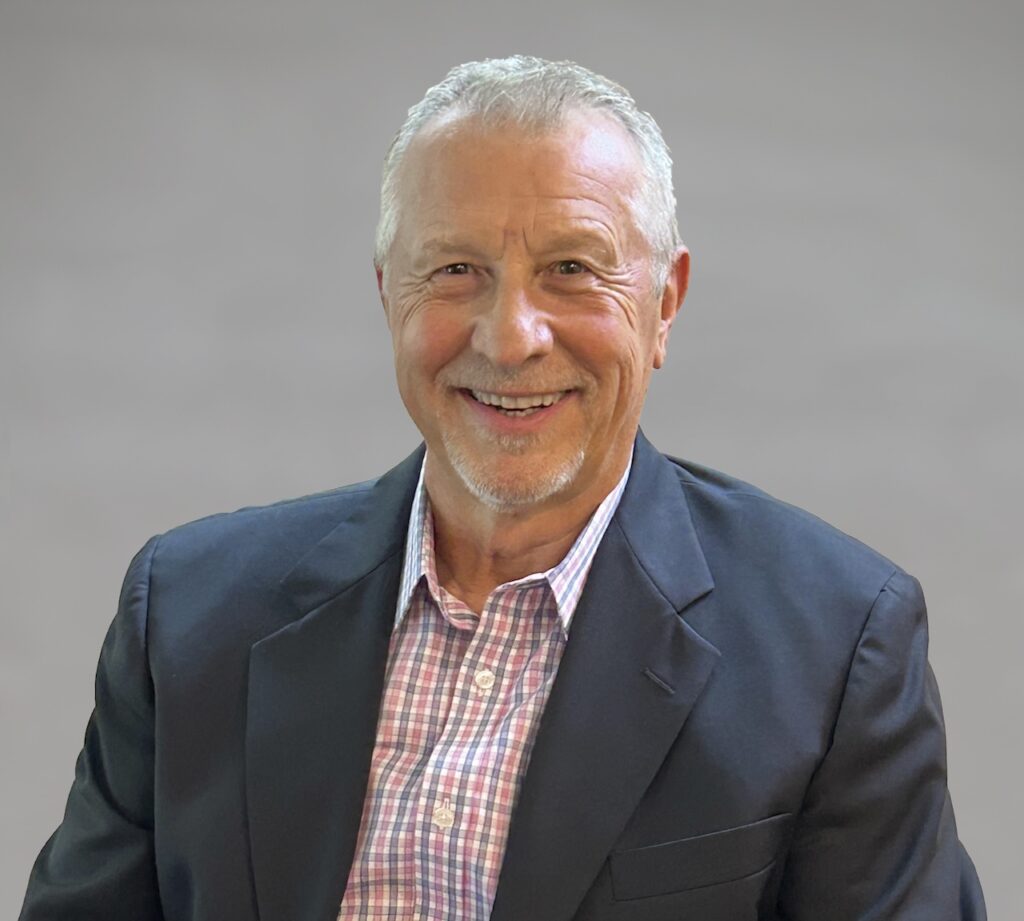 A smiling man with gray hair and a beard is wearing a dark blazer over a checkered shirt against a plain background.