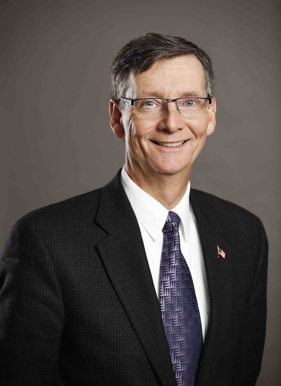 A person in a suit and tie, with short hair and glasses, smiles at the camera.