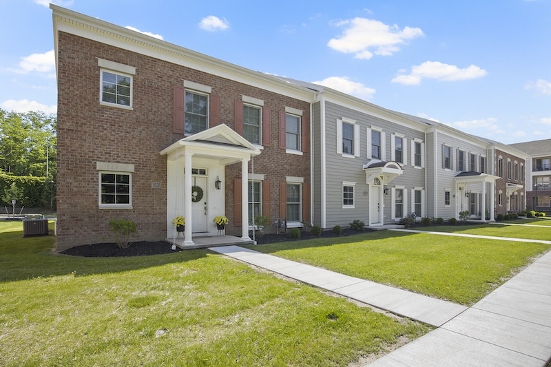 A row of two-story townhouses with brick and siding exteriors, small front porches, and manicured lawns under a blue sky.