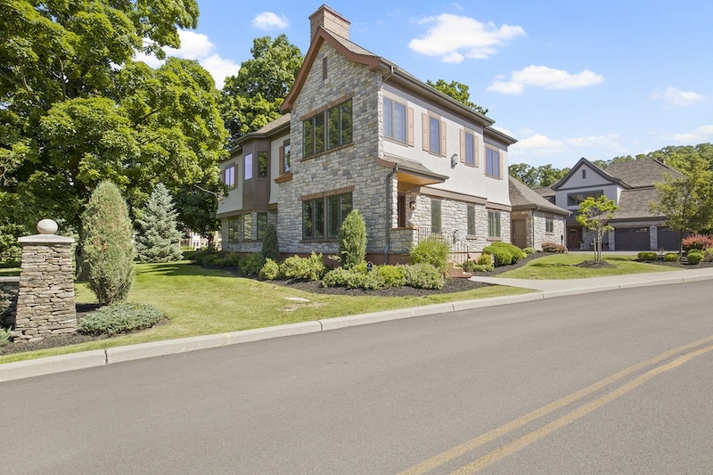 Two-story stone house with manicured landscaping and a paved street. Clear sky and trees in the background.