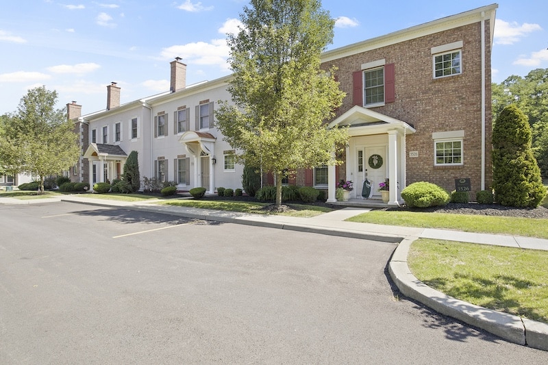 Row of two-story brick townhouses with neatly trimmed bushes, trees, and small front porches. Each unit features a gabled entrance and street parking is visible.