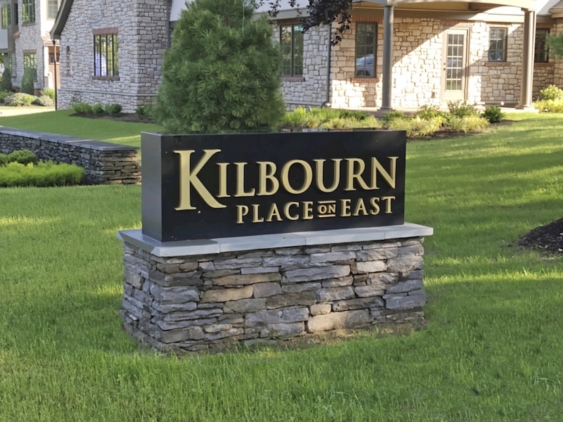 Sign for "Kilbourn Place on East" on a stone base, set in a grassy area near a stone building.