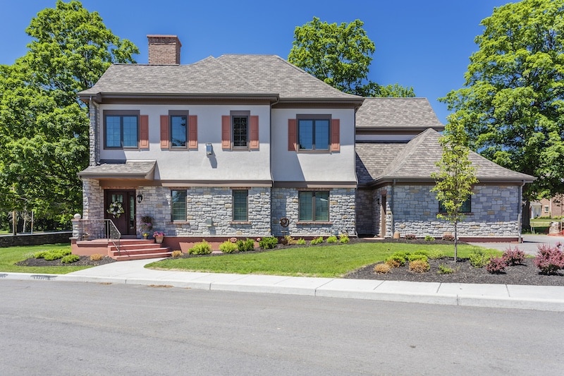 A two-story stone and stucco house with a chimney and brown accents, surrounded by trees and a manicured lawn on a clear day.