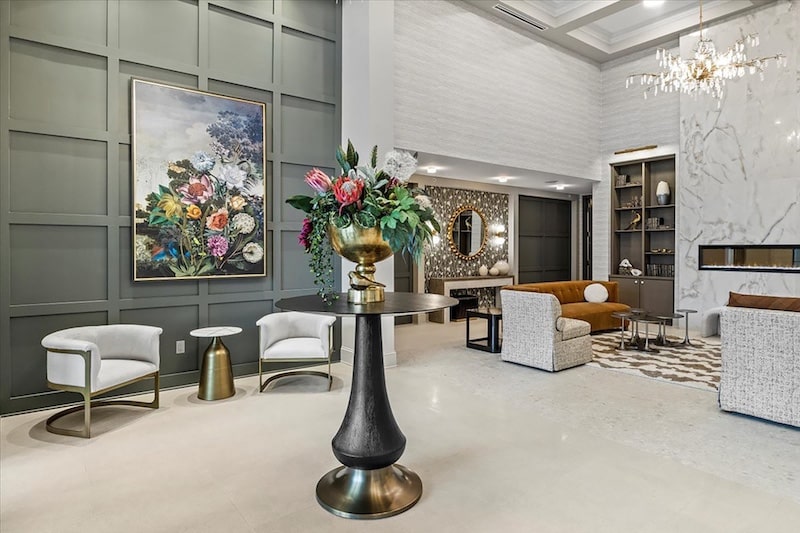 A modern lobby with elegant seating, a large floral painting, and a central round table with a flower arrangement. There's a chandelier above and a marble accent wall with built-in shelves.