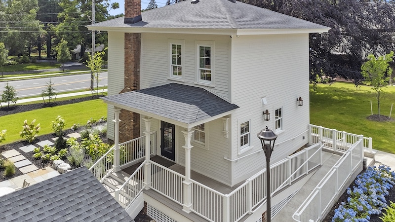 Two-story white house with a gabled roof, wraparound porch, and chimney. Surrounded by a landscaped garden with pathways and a street in the background.