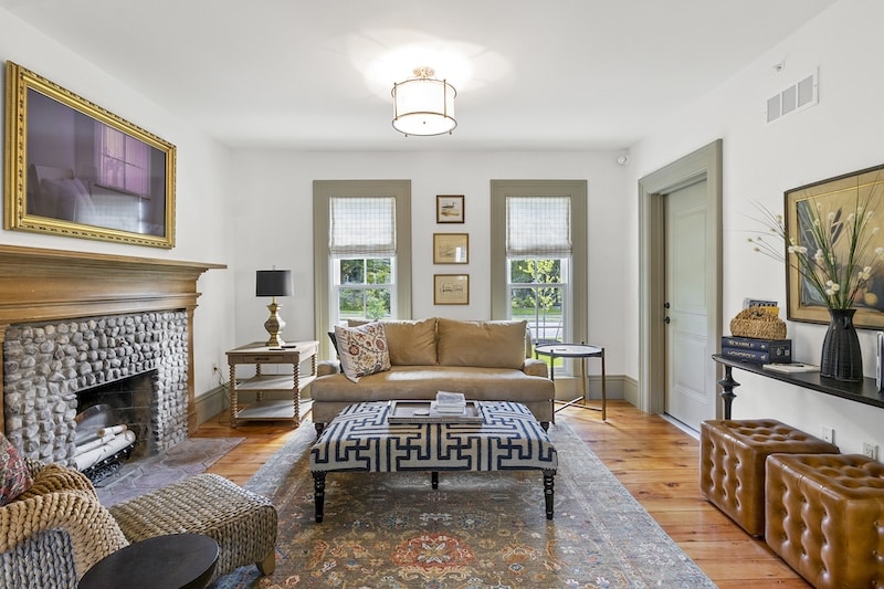 A cozy living room with a stone fireplace, beige couch, patterned ottoman, and decorative elements like framed art and vases. Natural light enters through two windows.