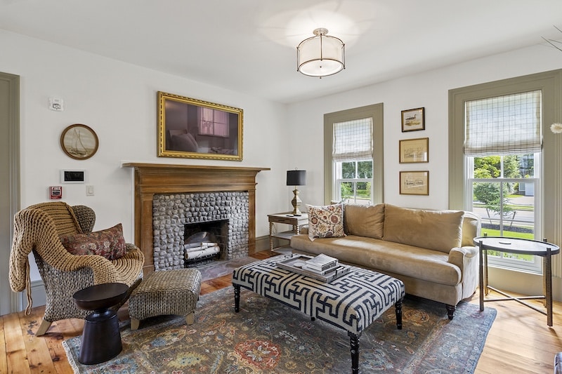 Cozy living room with a beige couch, patterned ottoman, and a wicker chair. Fireplace with a mirror above. Two windows allow natural light. Colorful rug on wooden floor.