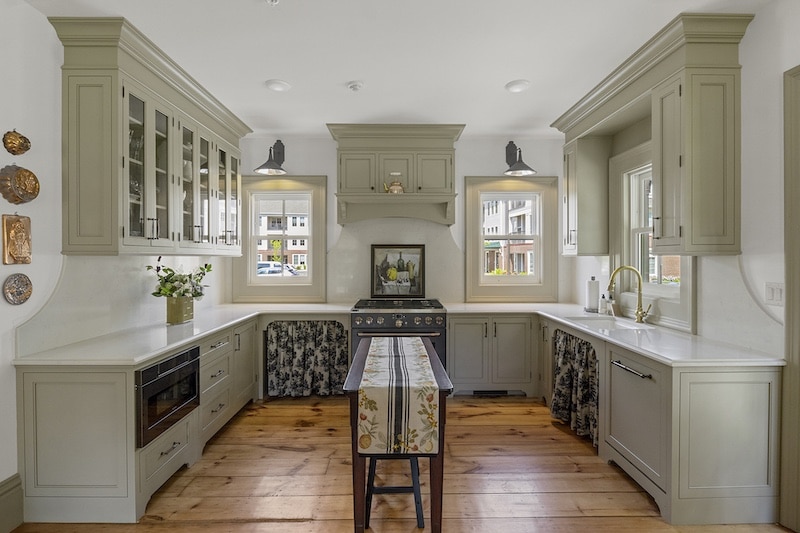 A kitchen with light gray cabinets, a central stove, and wooden floors. There are two windows, hanging lights, and floral-patterned curtains under the counters.