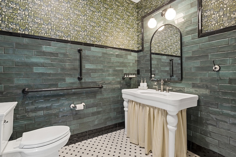 A bathroom with green tiled walls, a white sink with a skirt, an oval mirror, two lights above, and a white toilet on black-and-white patterned floor tiles.