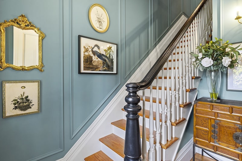 A staircase with wooden steps and white railings. The walls are painted blue and decorated with framed art. A wooden cabinet with a vase of flowers is on the right. A mirror hangs on the left.
