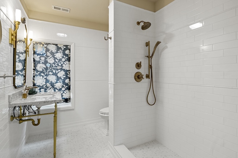 White tiled bathroom with a shower, gold fixtures, and a floral-patterned window covering.