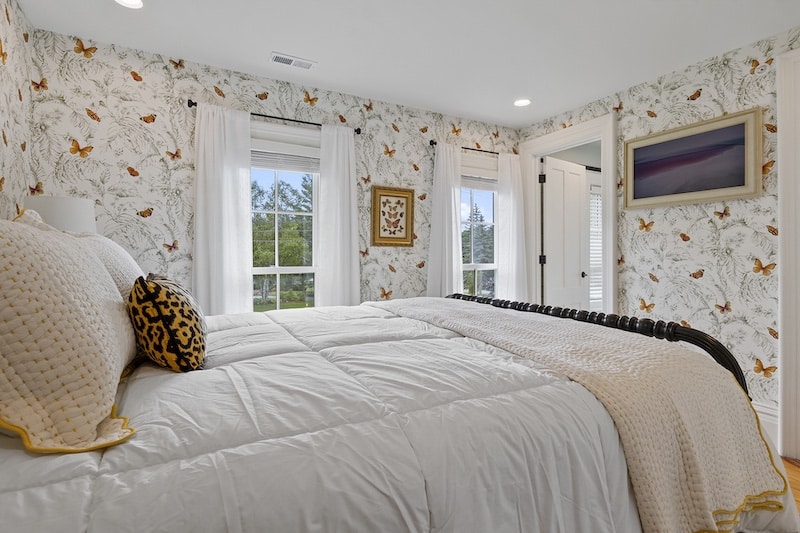Bedroom with floral wallpaper, featuring a large bed with white bedding and a decorative leopard print pillow. Windows with white curtains let in natural light, and a framed picture hangs on the wall.
