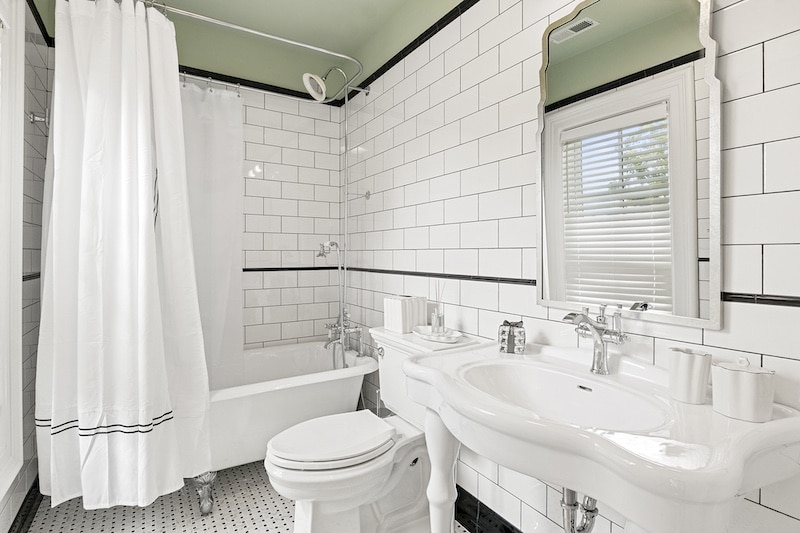 A white bathroom with a bathtub, shower curtain, toilet, and sink. The walls are tiled in white with black accents, and there is a mirror above the sink. A window with blinds is visible.