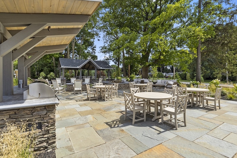 Outdoor patio area with round tables and chairs on stone flooring, shaded by trees. A wooden structure with a roof is in the background.