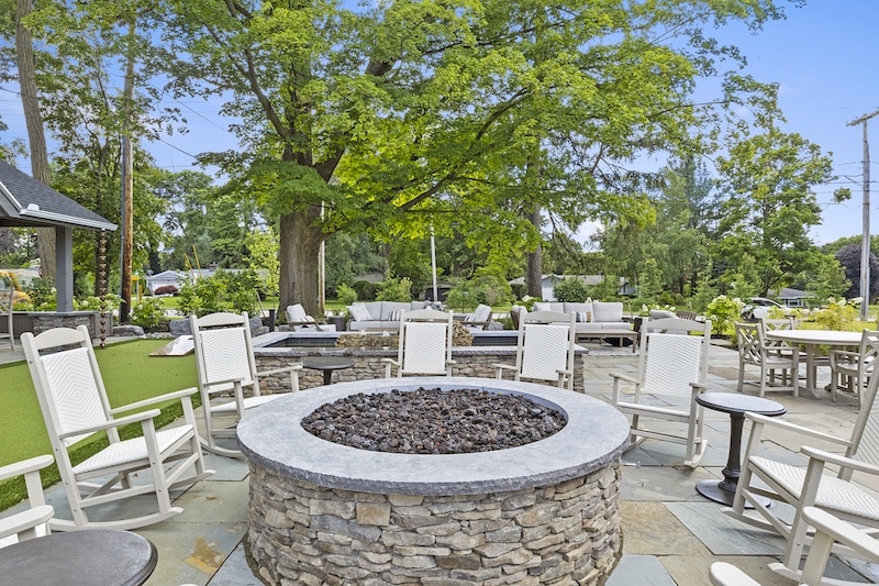 Outdoor patio area with stone fire pit surrounded by white rocking chairs, stone flooring, and green trees in the background.