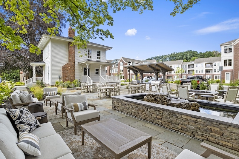 Outdoor patio area with seating, tables, and a stone water feature, surrounded by greenery and residential buildings in the background.