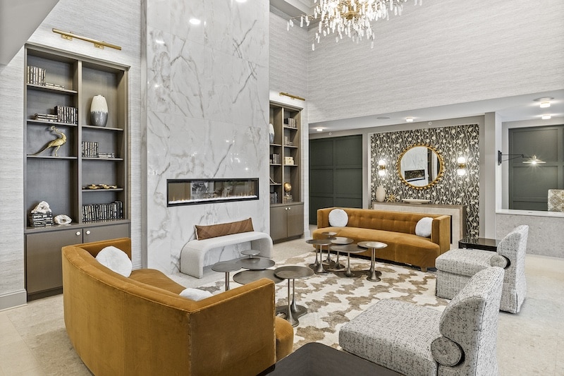 A modern lounge with marble fireplace, mustard couches, patterned chairs, decorative shelving, and a chandelier. A round mirror and abstract carpet complete the elegant setting.