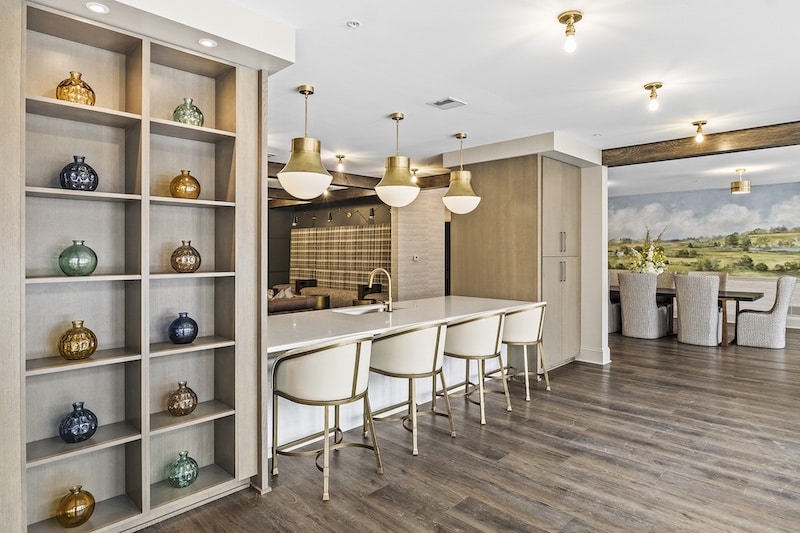 Modern kitchen with a white island, gold-accented chairs, open shelving with decorative vases, and pendant lights. A dining area with a landscape mural is visible in the background.