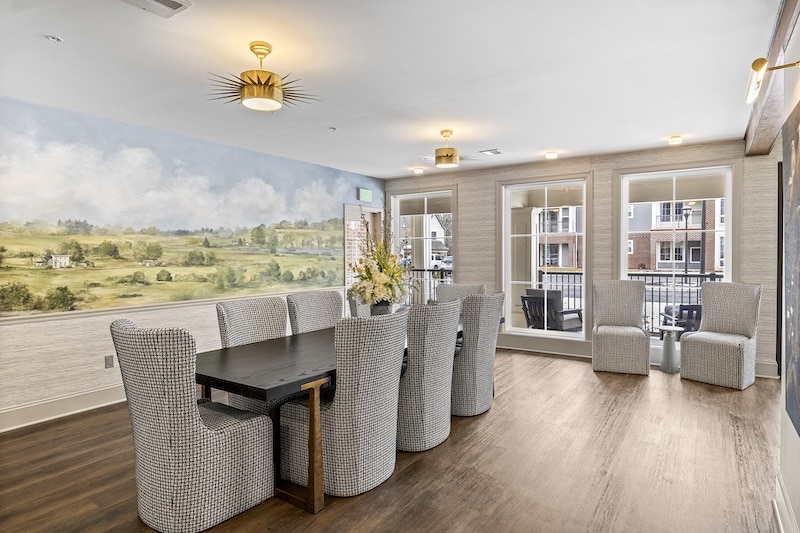 Dining room featuring a long dark table with patterned chairs, a mural of a landscape on the wall, and three sets of windows overlooking a balcony with additional seating.