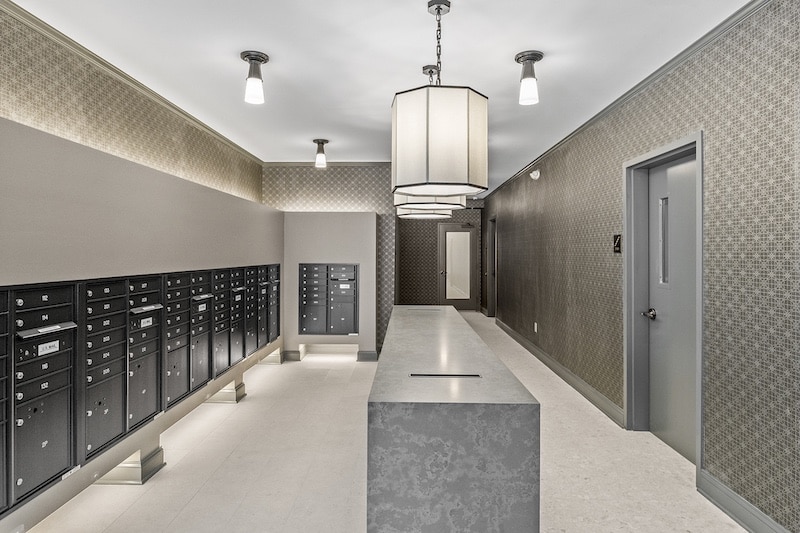 A modern mailroom with patterned walls and ceiling lights. Rows of black mailboxes on one side and a long table in the center.