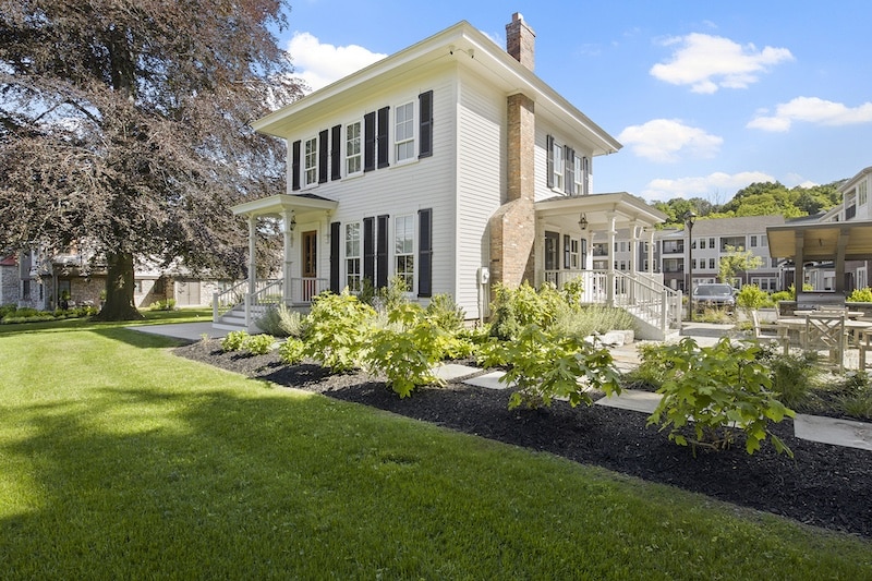 White two-story house with a porch, black shutters, and a brick chimney. Lush green lawn and landscaped bushes in the foreground. Clear blue sky with some clouds.