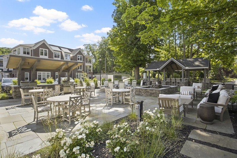Outdoor patio with tables and chairs surrounded by greenery, a pavilion, and a building in the background.