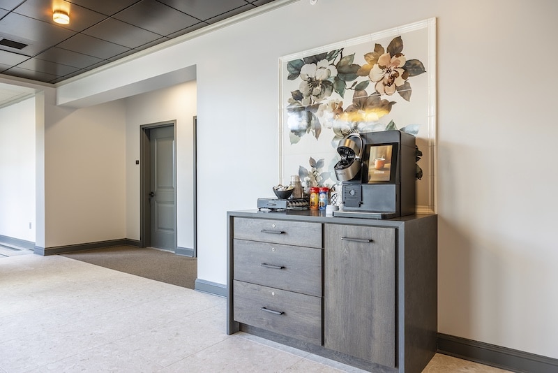 A coffee station with a coffee maker and condiments on a wooden cabinet. A large floral artwork hangs above.