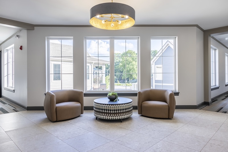 A modern lounge area with two beige armchairs and a patterned round table under a large ceiling light. Three windows in the background.