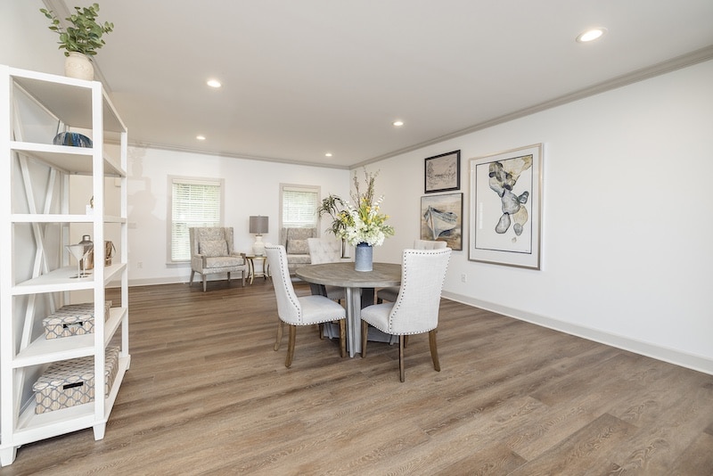 A modern living and dining area with wooden flooring, a round table with chairs, wall art, and large windows. Shelves on the left hold various decorative items.