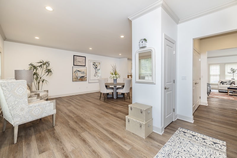 A spacious living area with wooden flooring, light-colored walls, a dining table, armchair, and decorative art on the wall. Boxes are stacked near an entrance to another room.