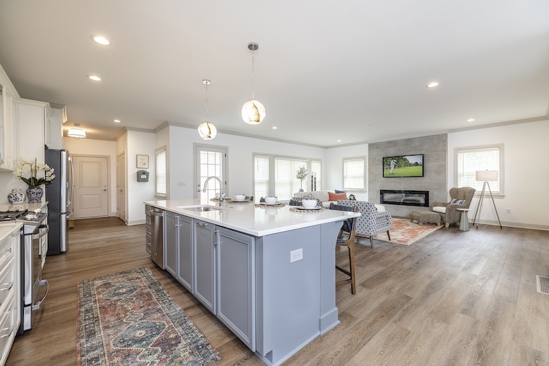 Modern open-concept living space with a kitchen island, pendant lighting, white cabinetry, and a seating area with a fireplace and wall-mounted TV. Wood flooring throughout.
