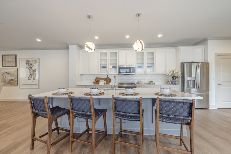 Modern kitchen with a white island, four wooden stools, pendant lights, stainless steel appliances, and white cabinetry.