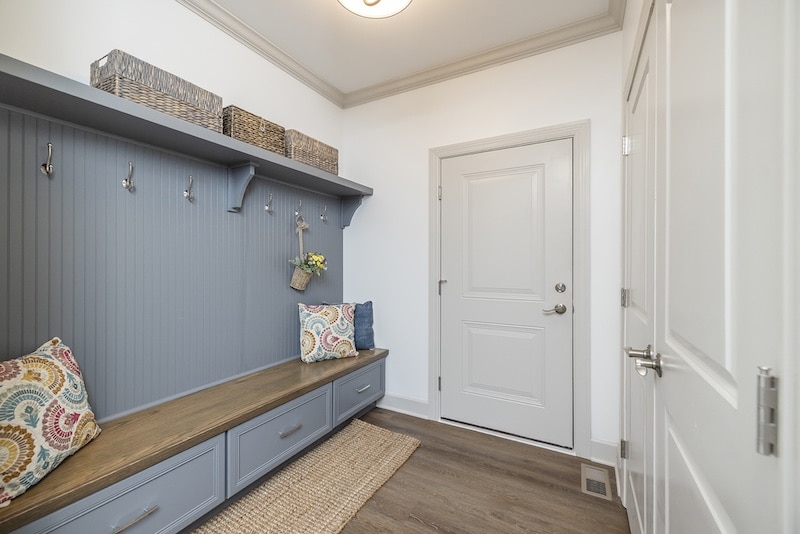 A mudroom with a bench, pillow, and storage drawers under coat hooks. Baskets are on the shelf above. A small plant hangs, and a patterned rug lies on the wooden floor.
