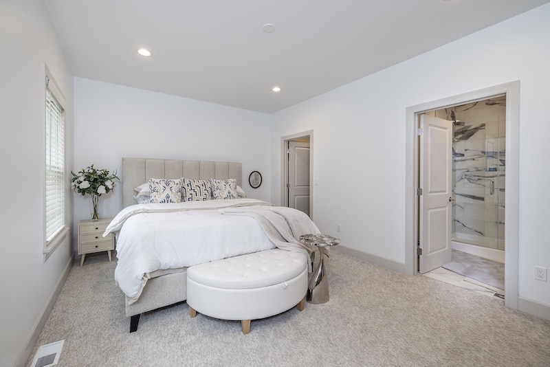 A modern bedroom with a gray upholstered bed, white bedding, a round white ottoman, two side tables, and an open door leading to a bathroom with marble accents.