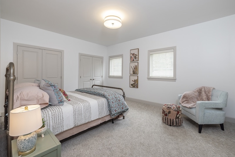 Bedroom with a metal bed, decorative pillows, a lamp on a nightstand, a light blue armchair, and framed artwork on a wall. Beige carpet and closed white doors are visible.