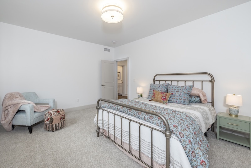 A bedroom with a metal bed frame, patterned bedding, two nightstands with lamps, a blue armchair, and a pouf on a carpeted floor. Walls and ceiling are white with a round light fixture.